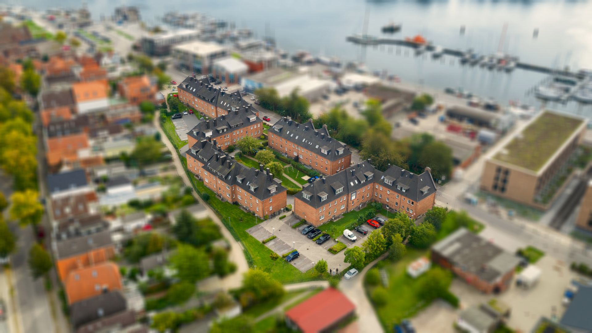 Mehrere rote Wohngebäude mit Innenhof und Parkplätzen, im Hintergrund ein Hafen.