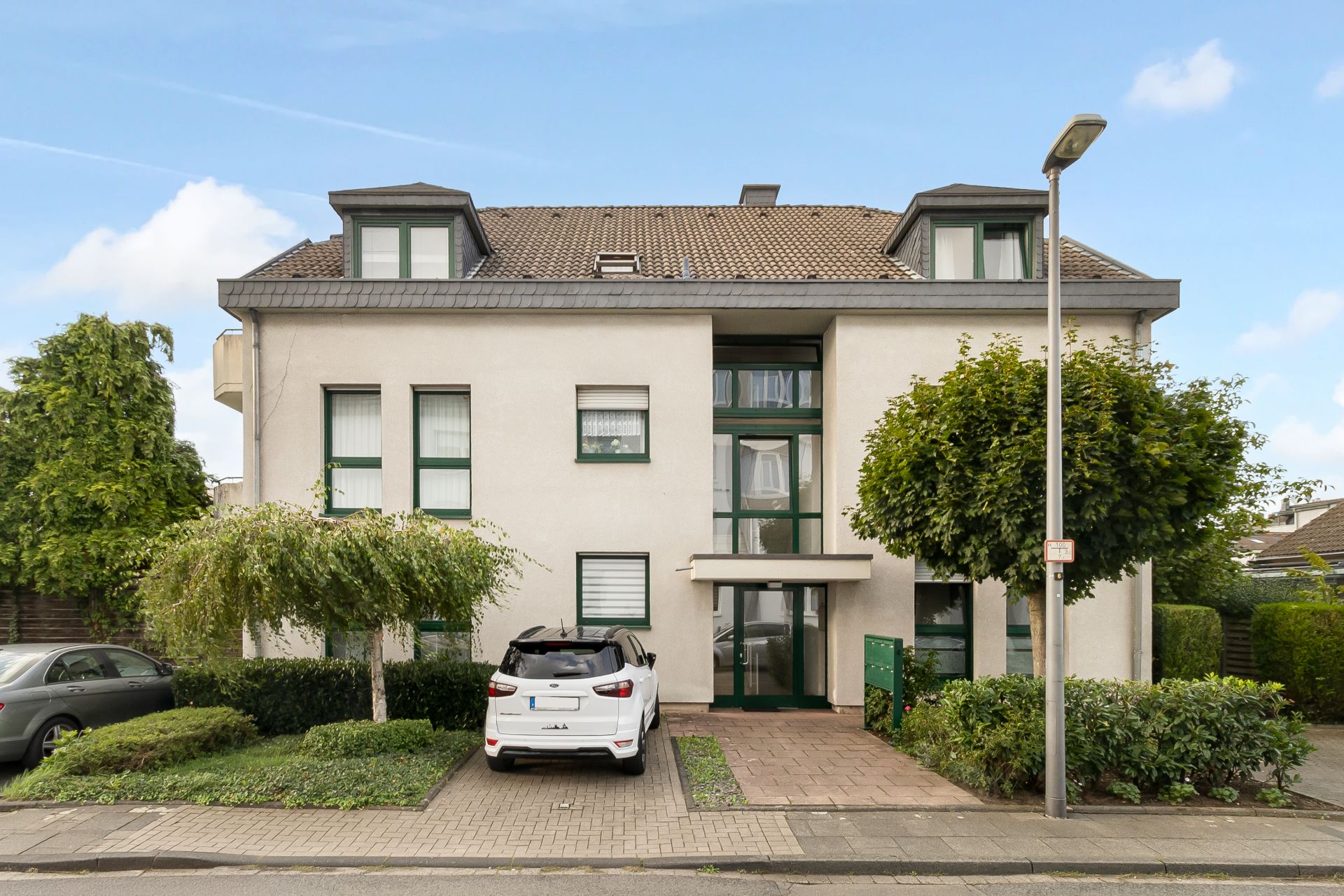 Zweistöckiges Wohnhaus mit Dachgauben, Bäumen vor dem Eingang und geparkten Autos davor.