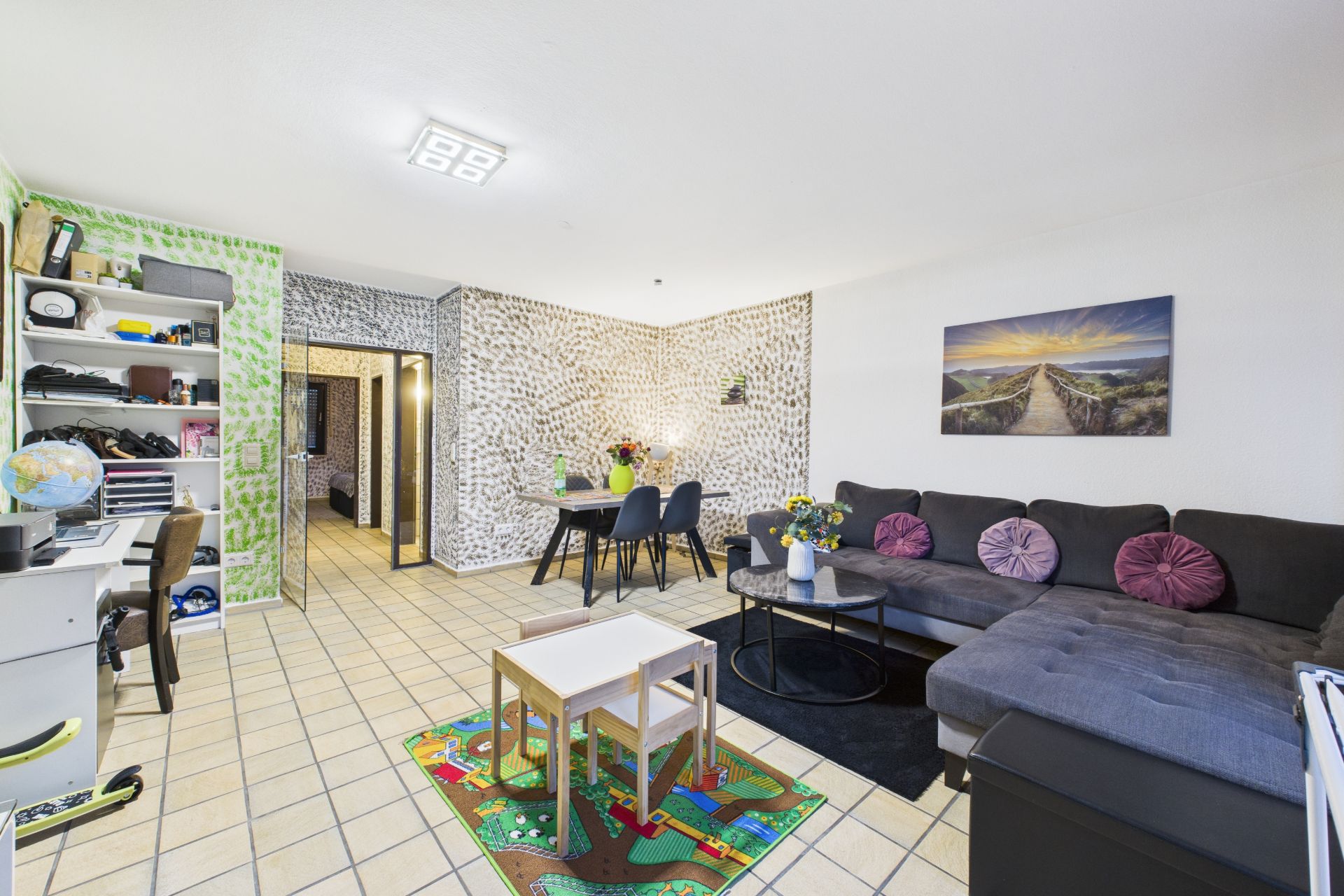 Helles Wohnzimmer mit Ecksofa, Esstisch, Spielteppich und Schreibtisch.