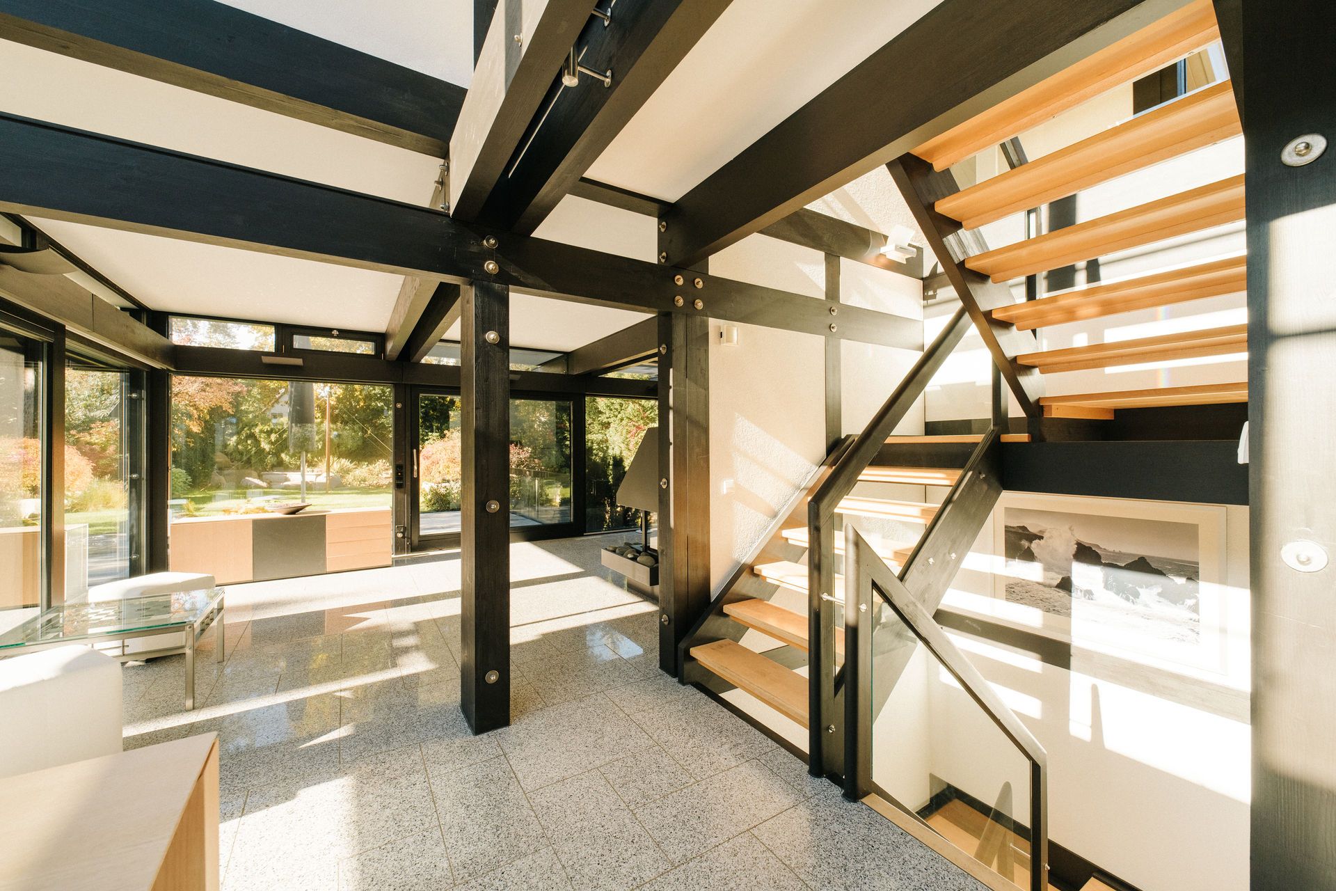 Heller Wohnbereich mit großen Fenstern, sichtbaren Stahlträgern und moderner Holztreppe.