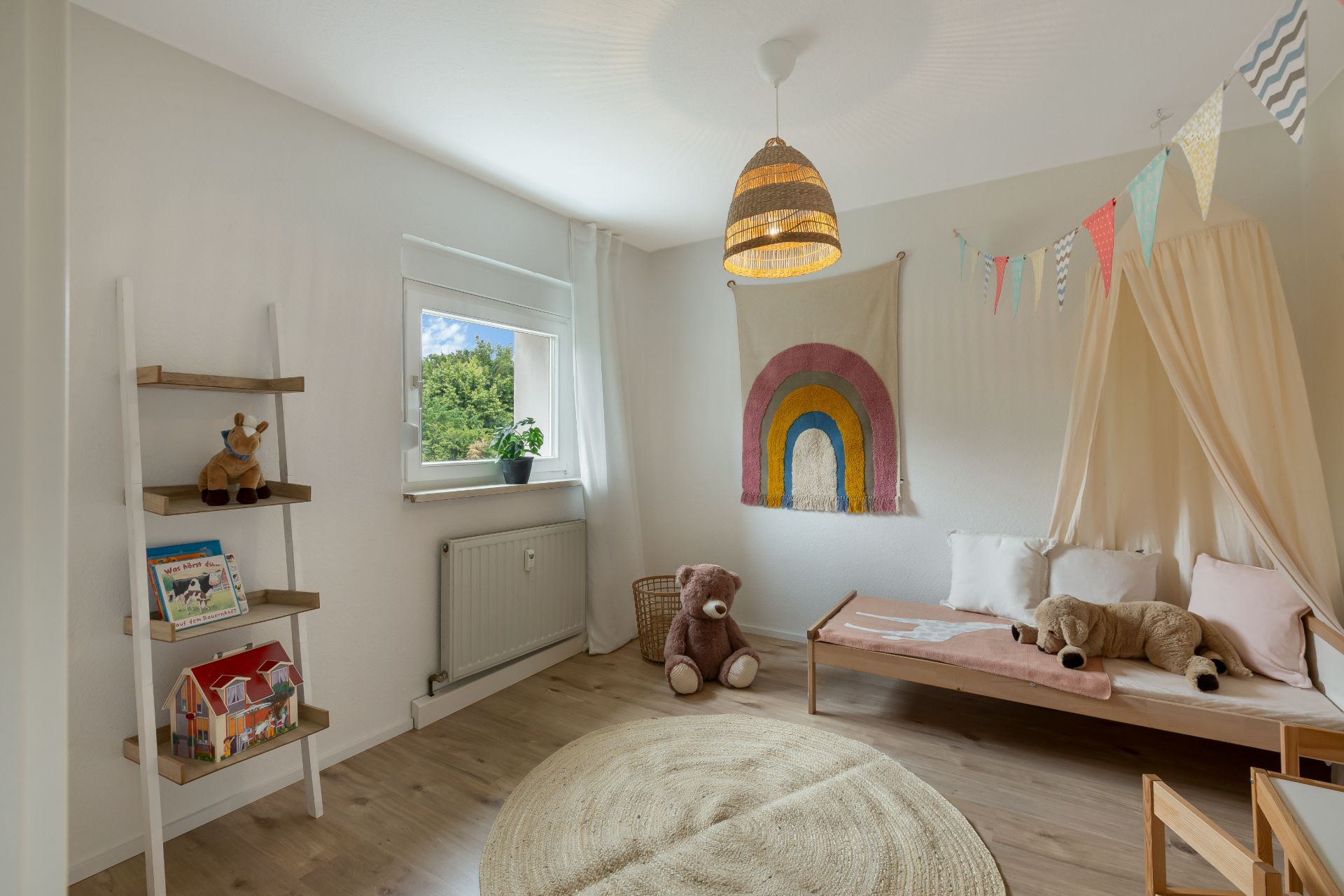 Helles Kinderzimmer mit Bett, Teppich, Kuscheltieren, Regal und Regenbogen-Wandbild.