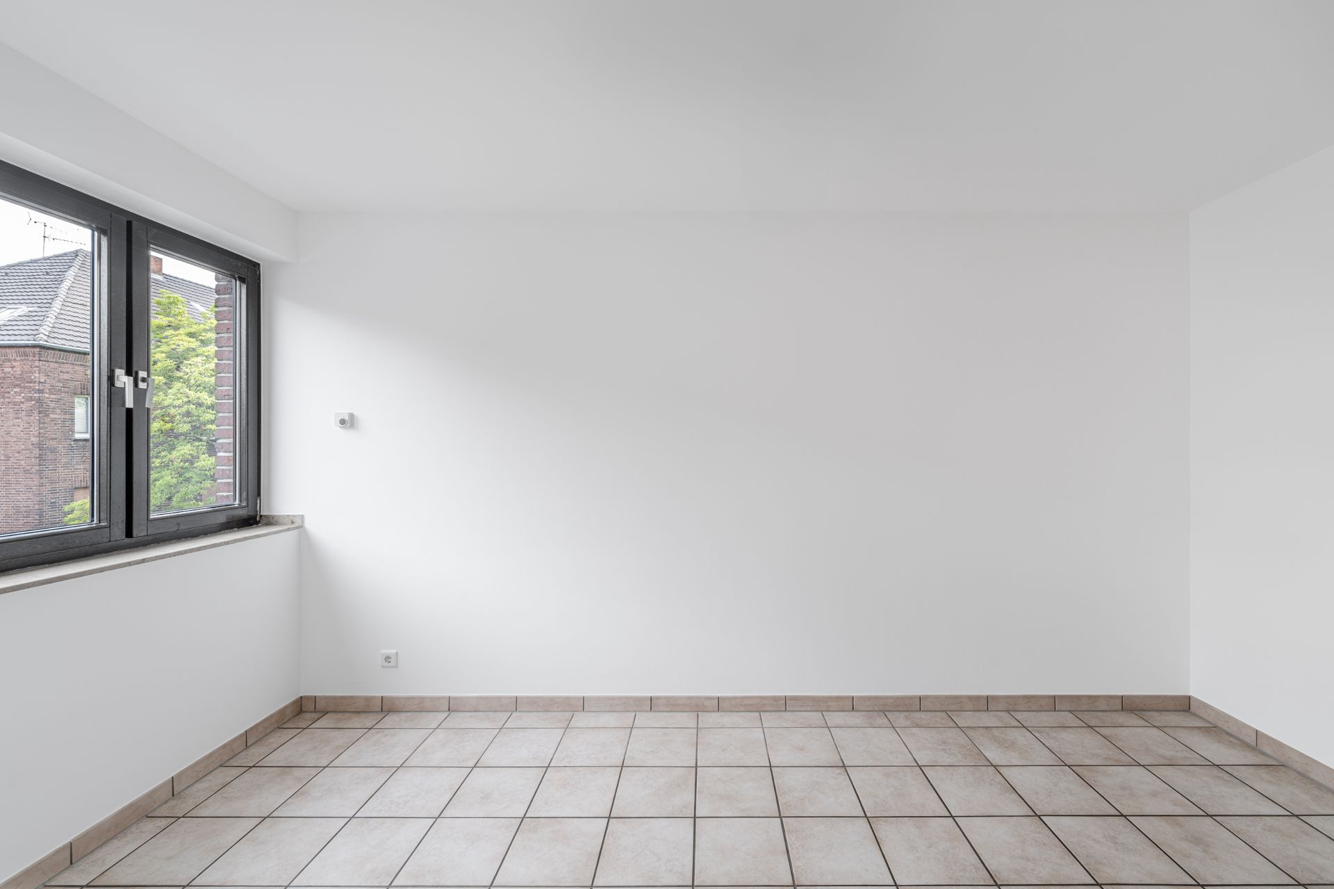 Leerer, heller Raum mit weißen Wänden, gefliestem Boden und großem Fenster.