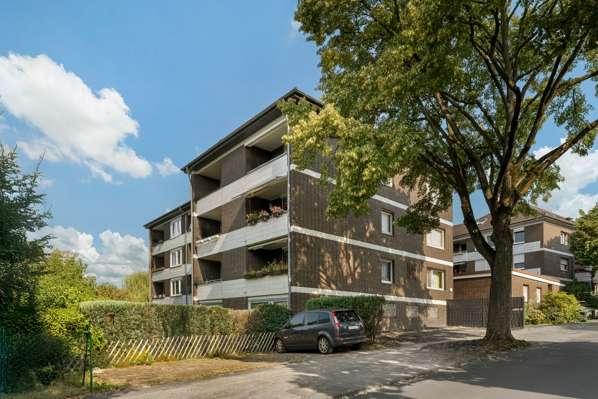 Mehrfamilienhaus mit Balkonen, umgeben von Bäumen und geparktem Auto davor.