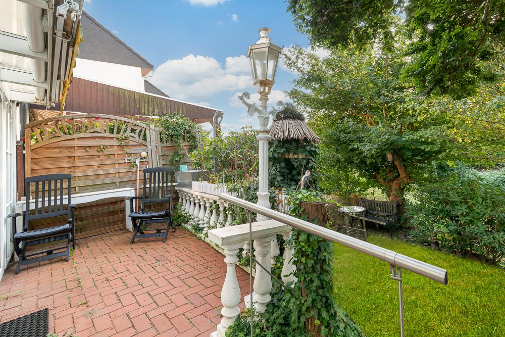 Terrasse mit zwei schwarzen Stühlen, Laterne und Blick auf grünen Garten mit Bäumen.