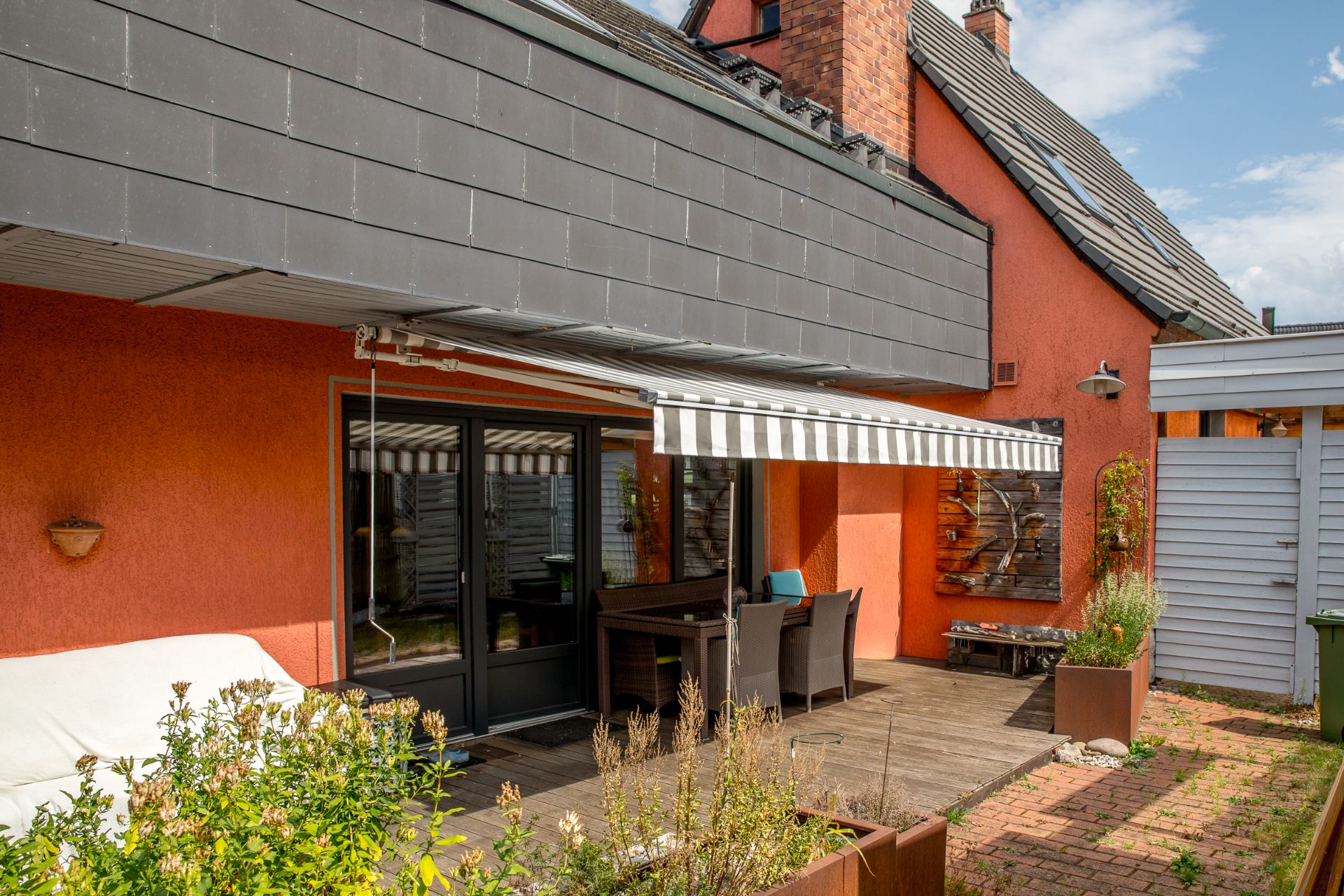 Terrasse mit Markise, Gartentisch, Stühlen und Pflanzkübeln an einem orangefarbenen Haus.