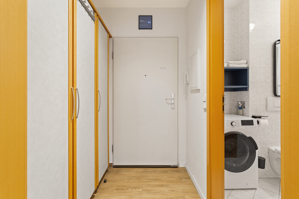 Flur mit weißer Eingangstür, Einbauschränken und Blick ins Badezimmer mit Waschmaschine.