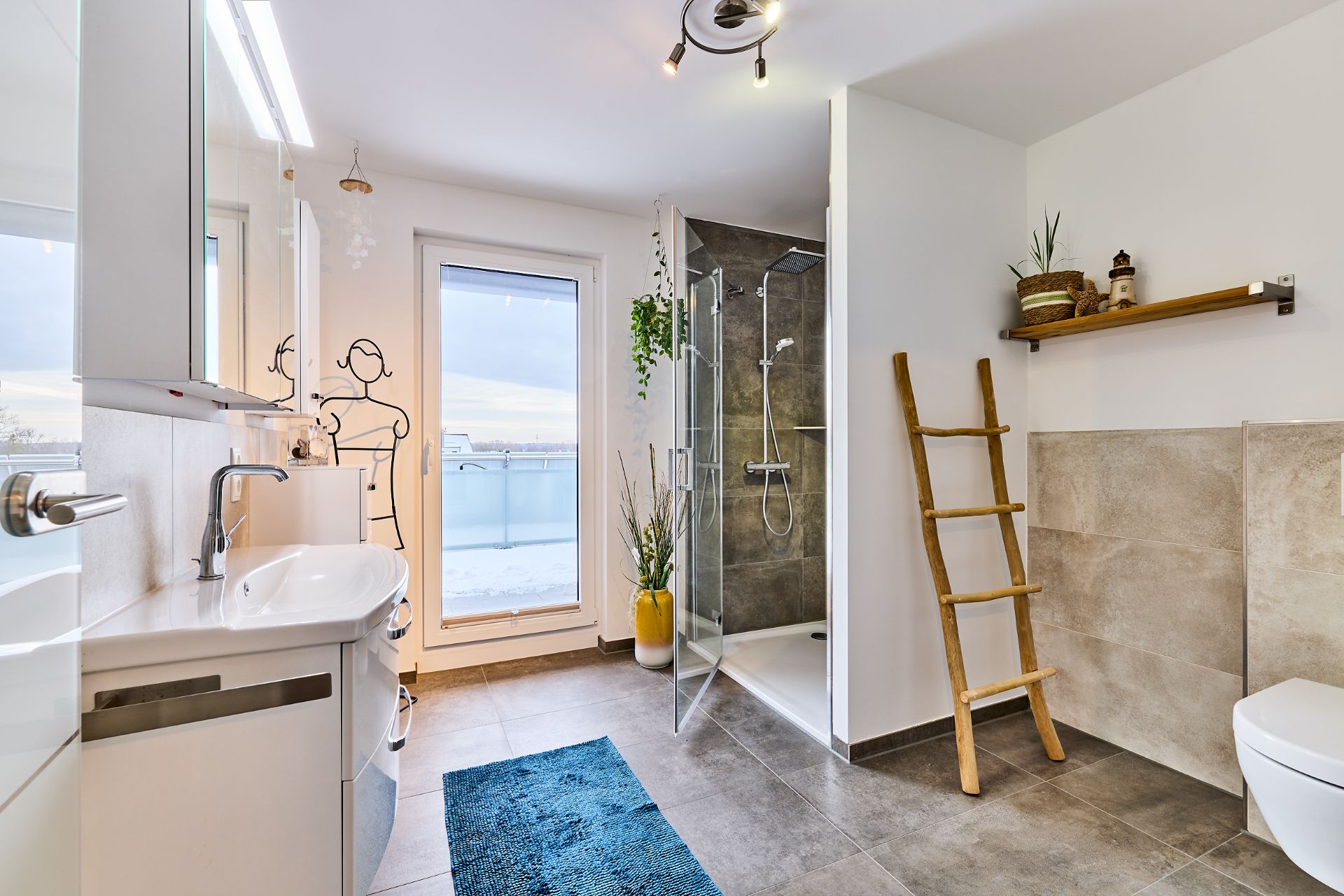 Modernes Badezimmer mit Dusche, Spiegel, Leiterregal, blauem Teppich und Zugang zum Balkon.