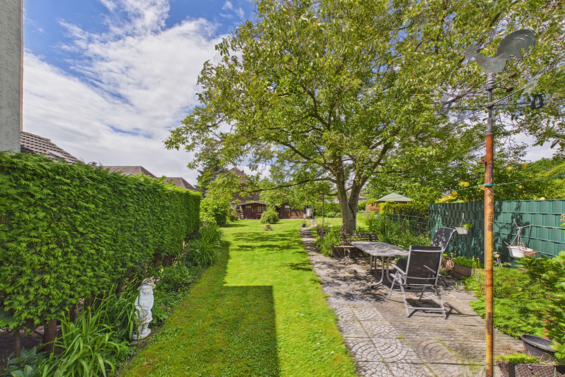 Grüner Garten mit Terrasse, Tisch und Stühlen, umgeben von Bäumen und Hecken.