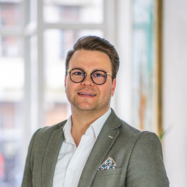 Ein Mann mit Brille und Sakko steht in einem hellen Raum mit Fenster im Hintergrund.