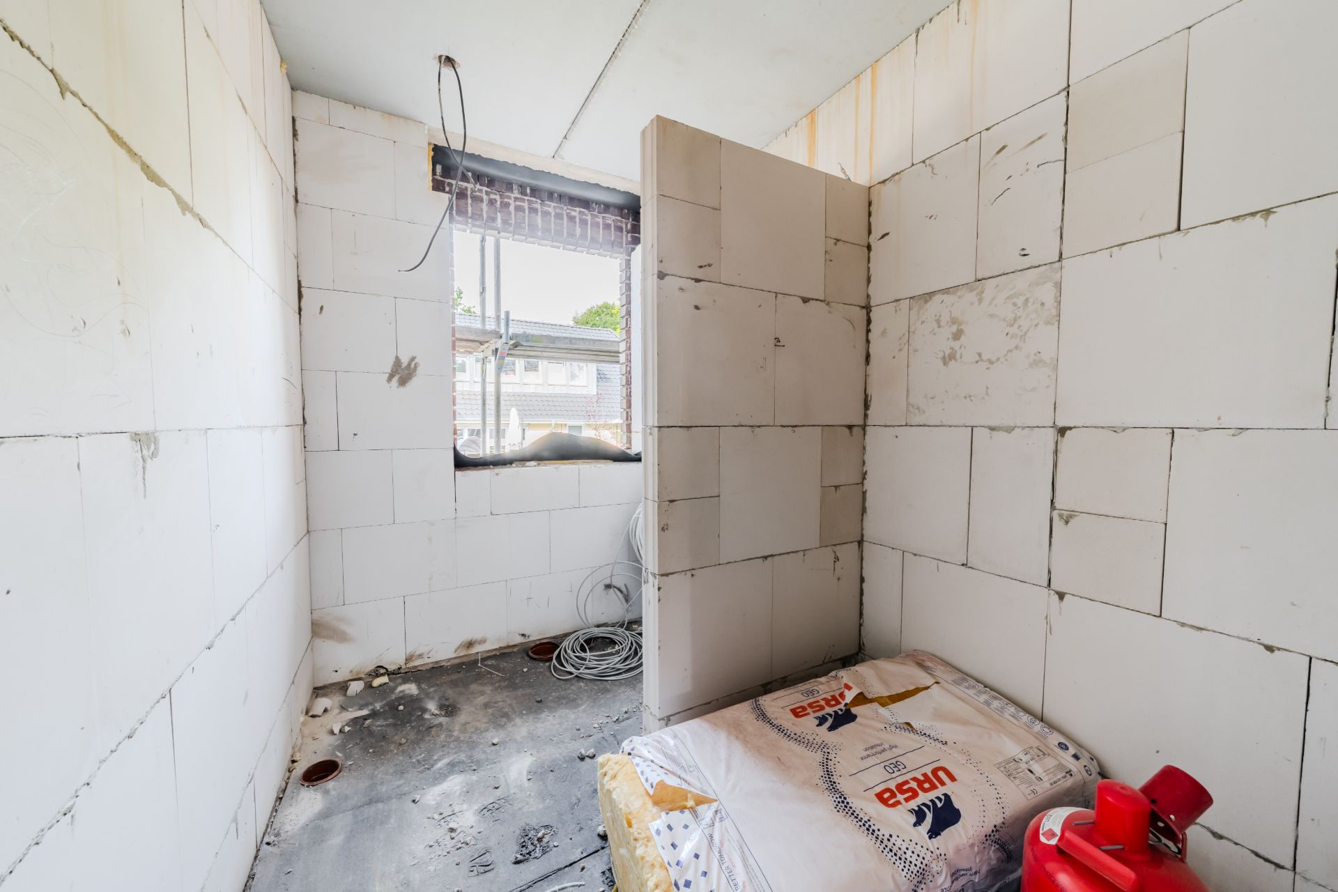 Kleiner Raum im Rohbau mit offenem Fenster, Baumaterial und Kabeln auf dem Boden.