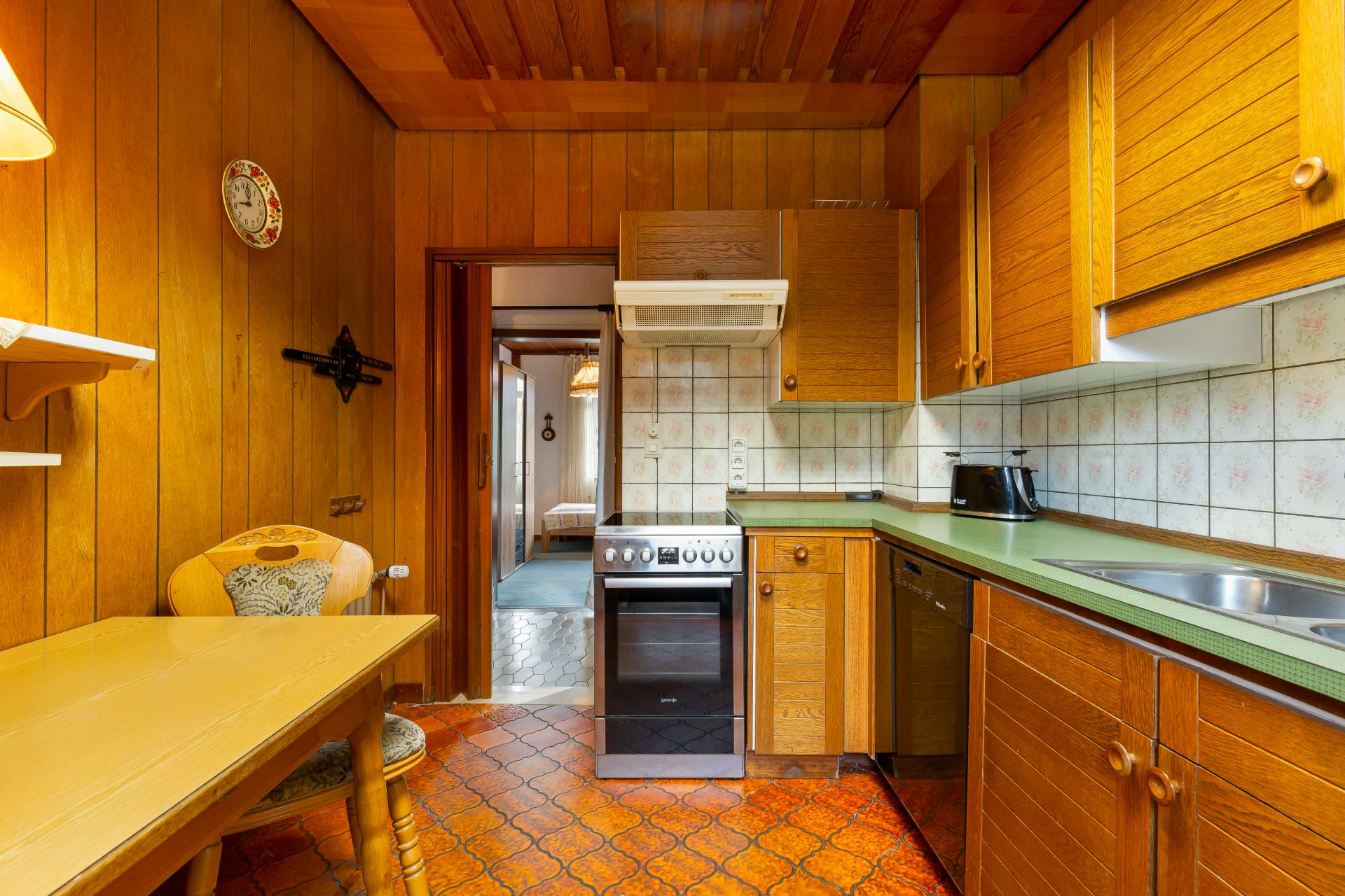 Holzgetäfelte Küche mit Herd, Spüle, Toaster, Tisch und zwei Stühlen, angrenzender Flur sichtbar.