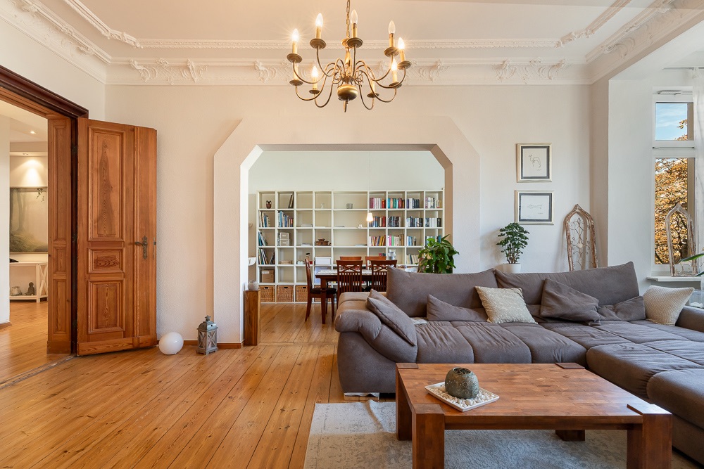 Wohnzimmer mit grauem Ecksofa, Holztisch, Kronleuchter und Blick auf Esszimmer mit Bücherregal.