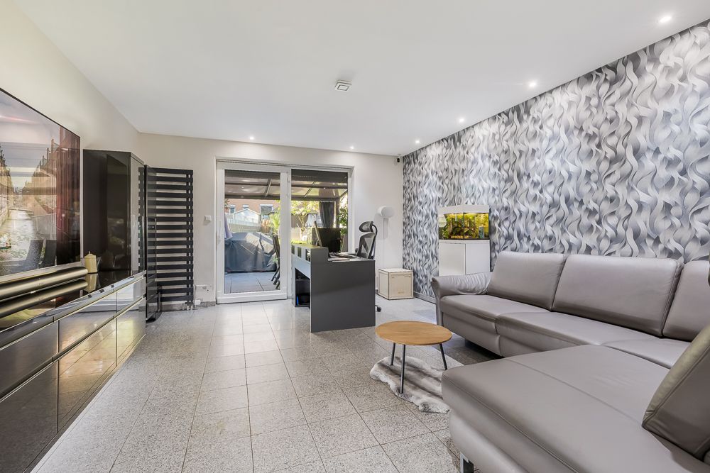 Modernes Wohnzimmer mit grauem Sofa, Schreibtisch, Aquarium und Glastüren zum Garten.