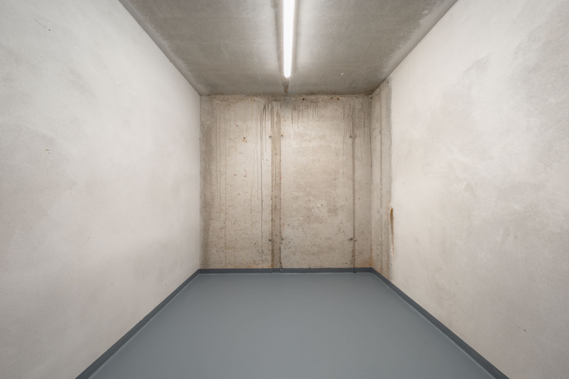 Kleiner leerer Raum mit Betonwänden, grauem Boden und Deckenleuchte.