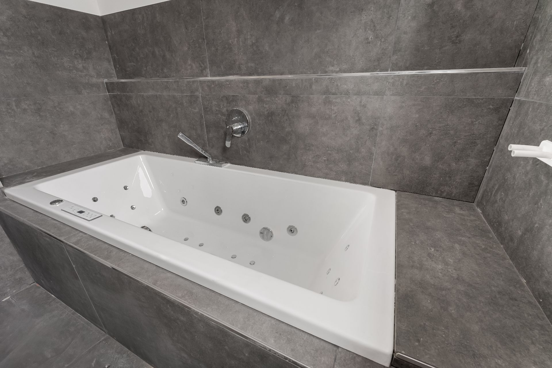 Weiße Eckbadewanne mit Whirlpooldüsen in einem Bad mit grauen Fliesen.