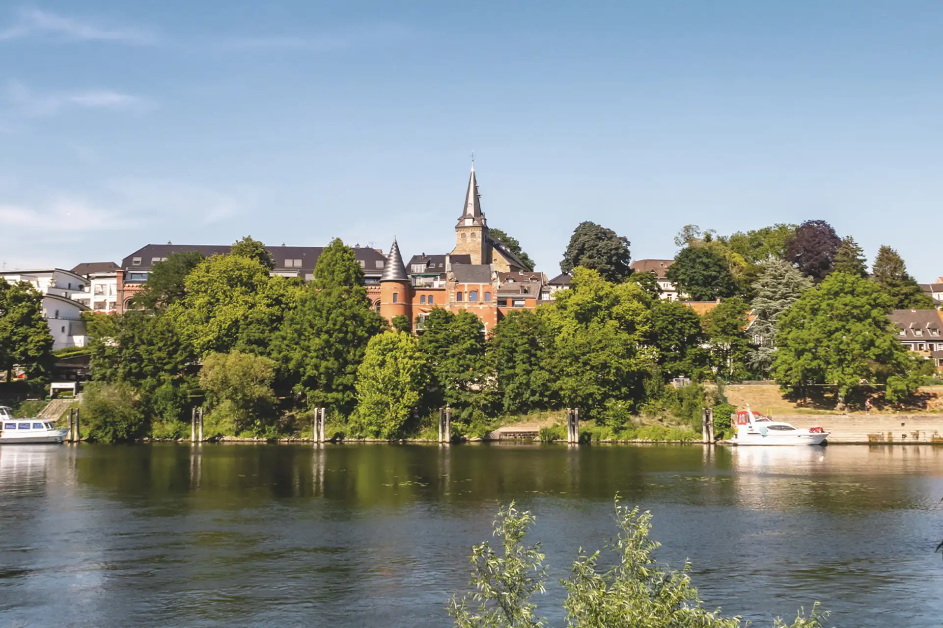 Stadtansicht mit Fluss, Bäumen und Gebäuden im Hintergrund bei klarem Himmel.