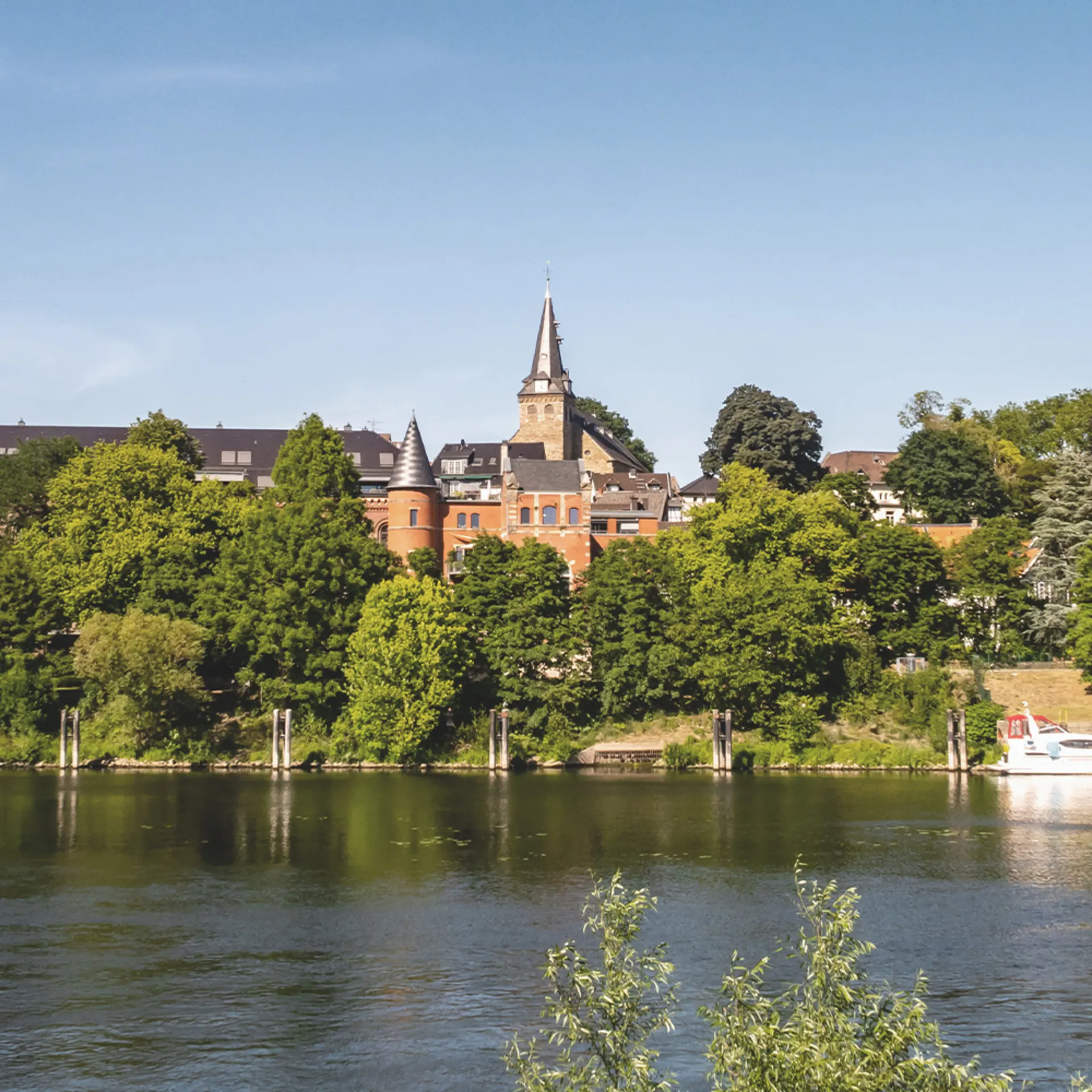 Stadtansicht mit Fluss, Bäumen und Gebäuden im Hintergrund bei klarem Himmel.