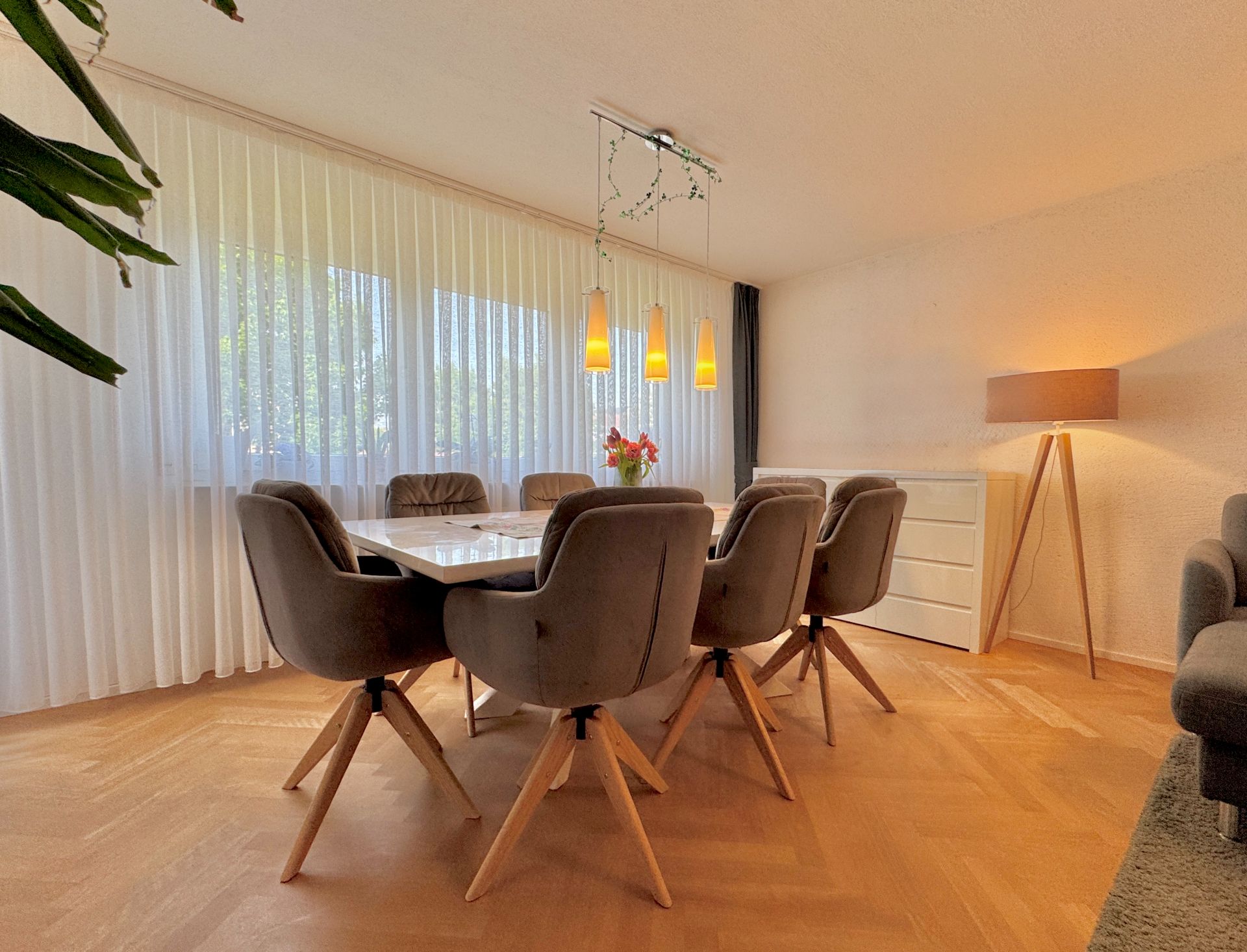 Esszimmer mit sechs grauen Stühlen, großem Tisch, Stehlampe und Fenster mit weißen Vorhängen.