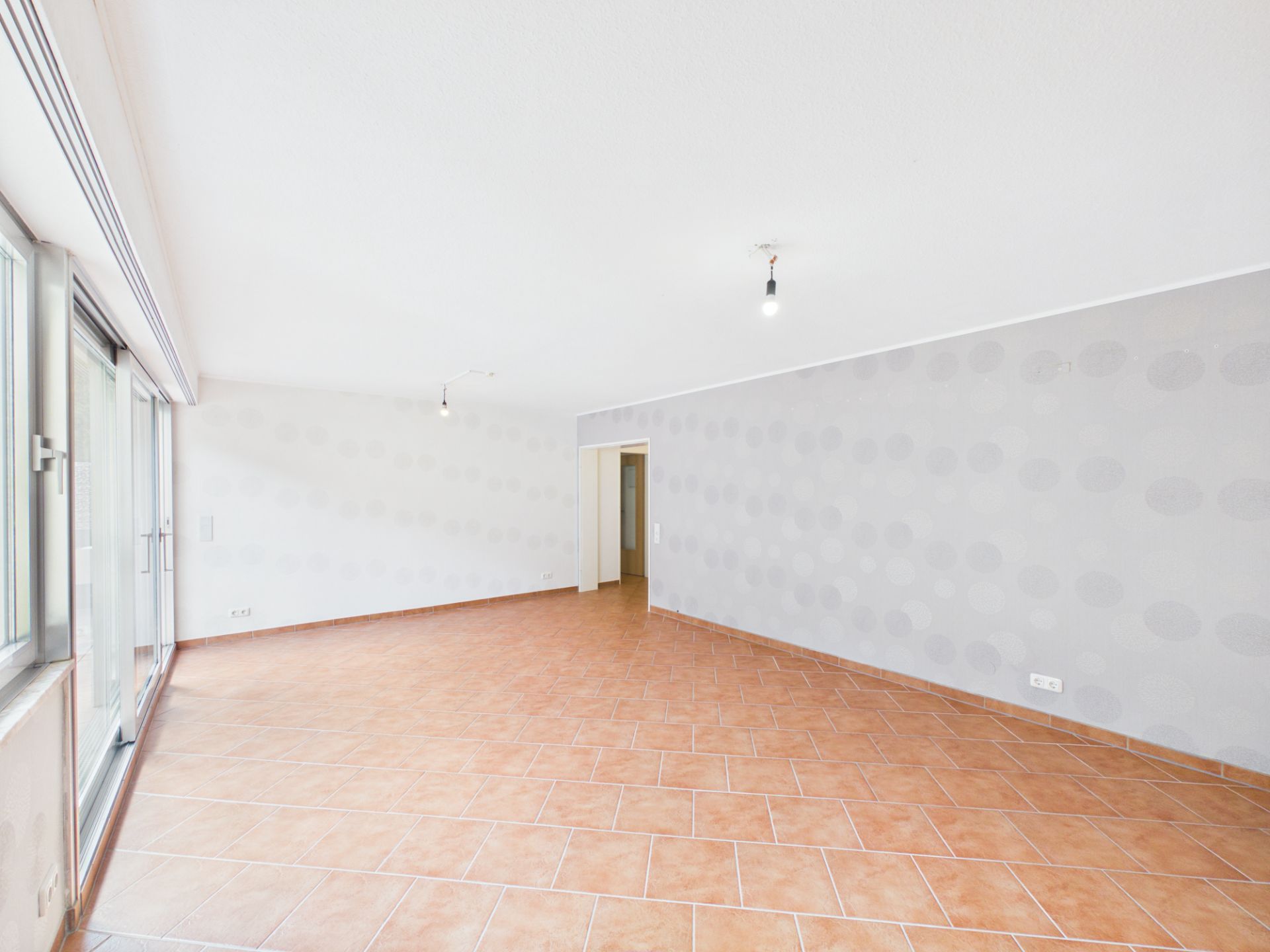 Heller leerer Raum mit Fliesenboden, großen Fenstern und einer Tür in der Rückwand.