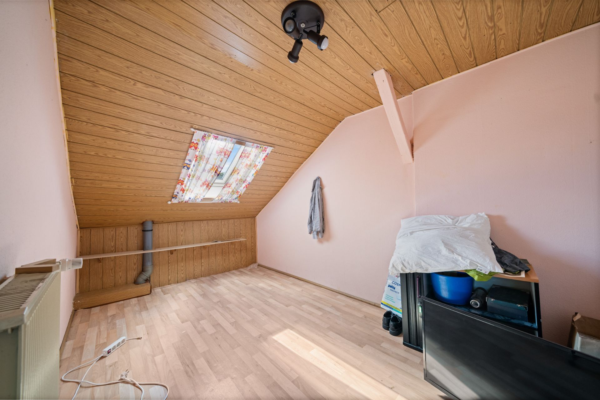Dachzimmer mit Holzdecke, schrägem Fenster und etwas Hausrat in einer Ecke.