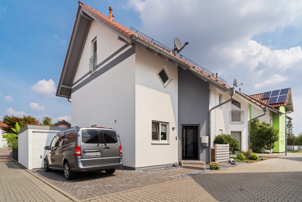Modernes Einfamilienhaus mit grau-weißer Fassade, geparktem Auto und kleinem Garten vor dem Eingang.