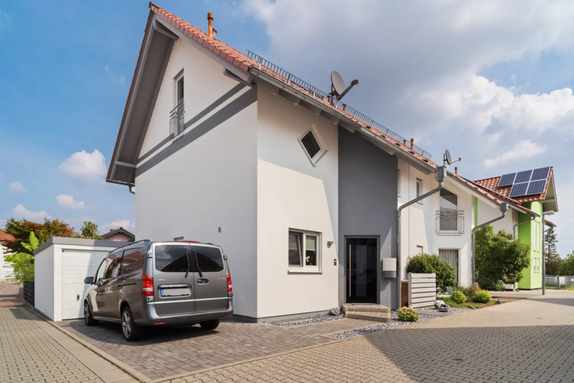 Modernes Einfamilienhaus mit grau-weißer Fassade, geparktem Auto und kleinem Garten vor dem Eingang.