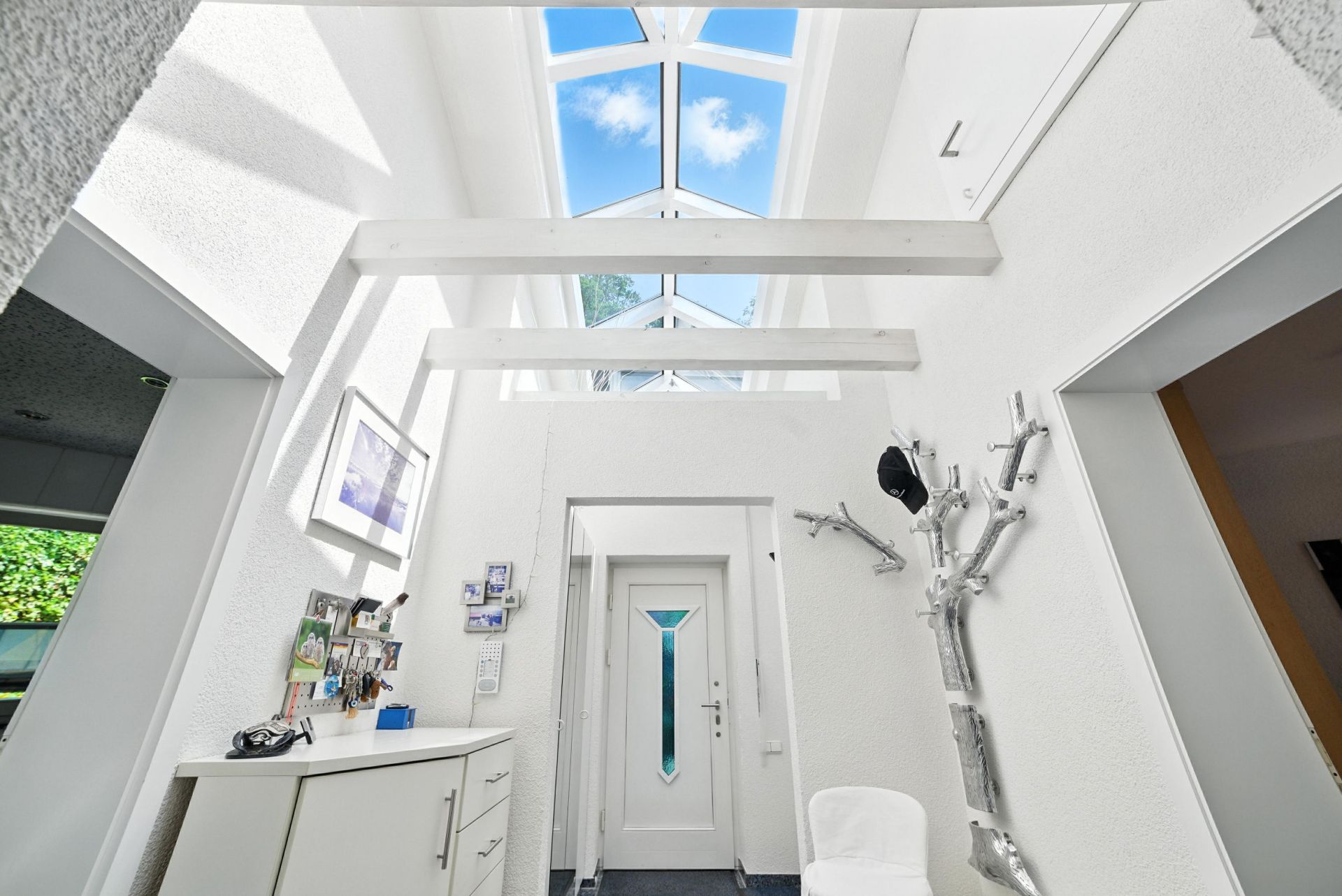 Heller Flur mit Glasdach, weißer Tür, Kommode und Garderobe in Baumform.