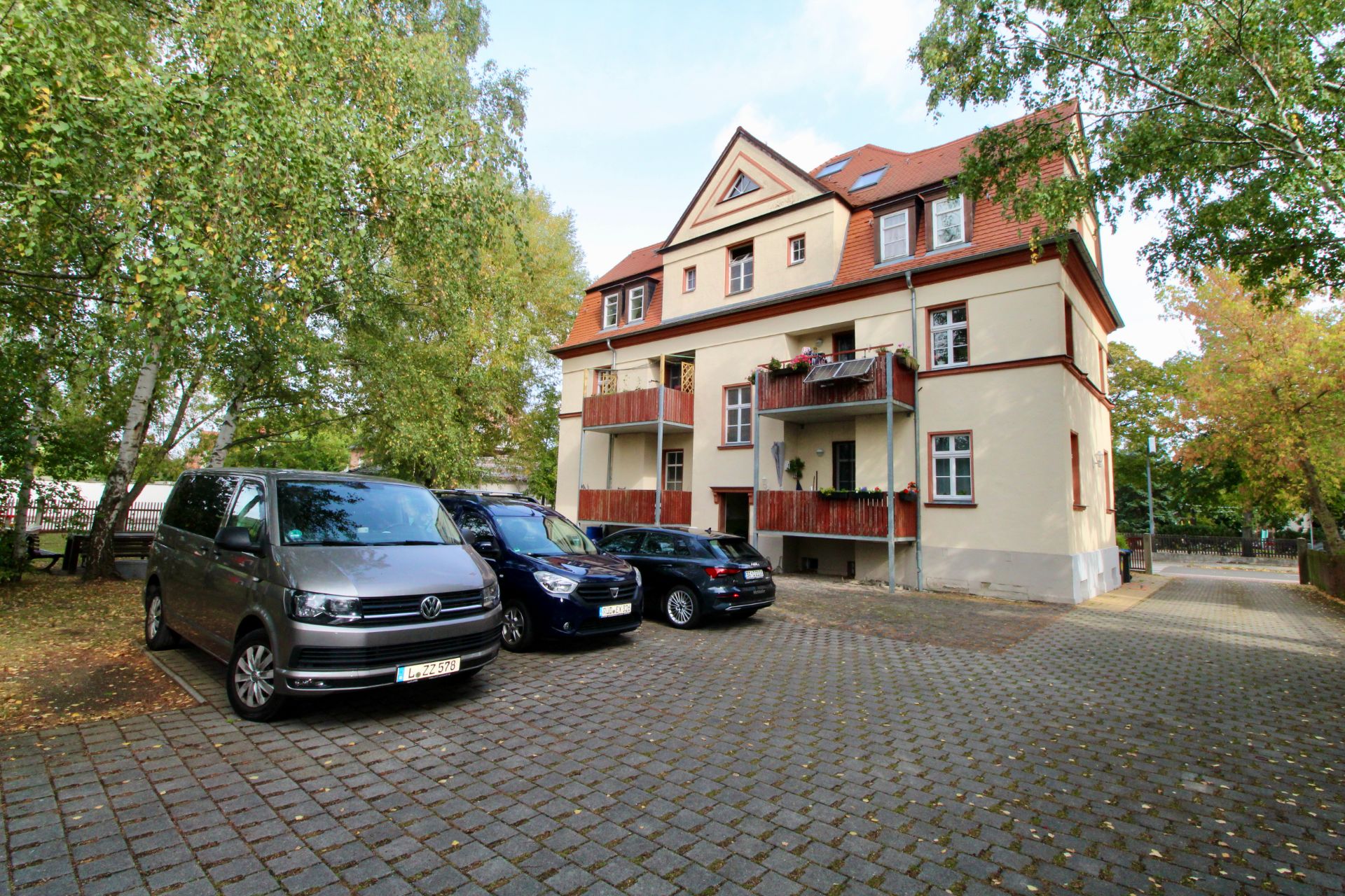 Mehrfamilienhaus mit rotem Dach und mehreren Autos auf gepflastertem Parkplatz davor.