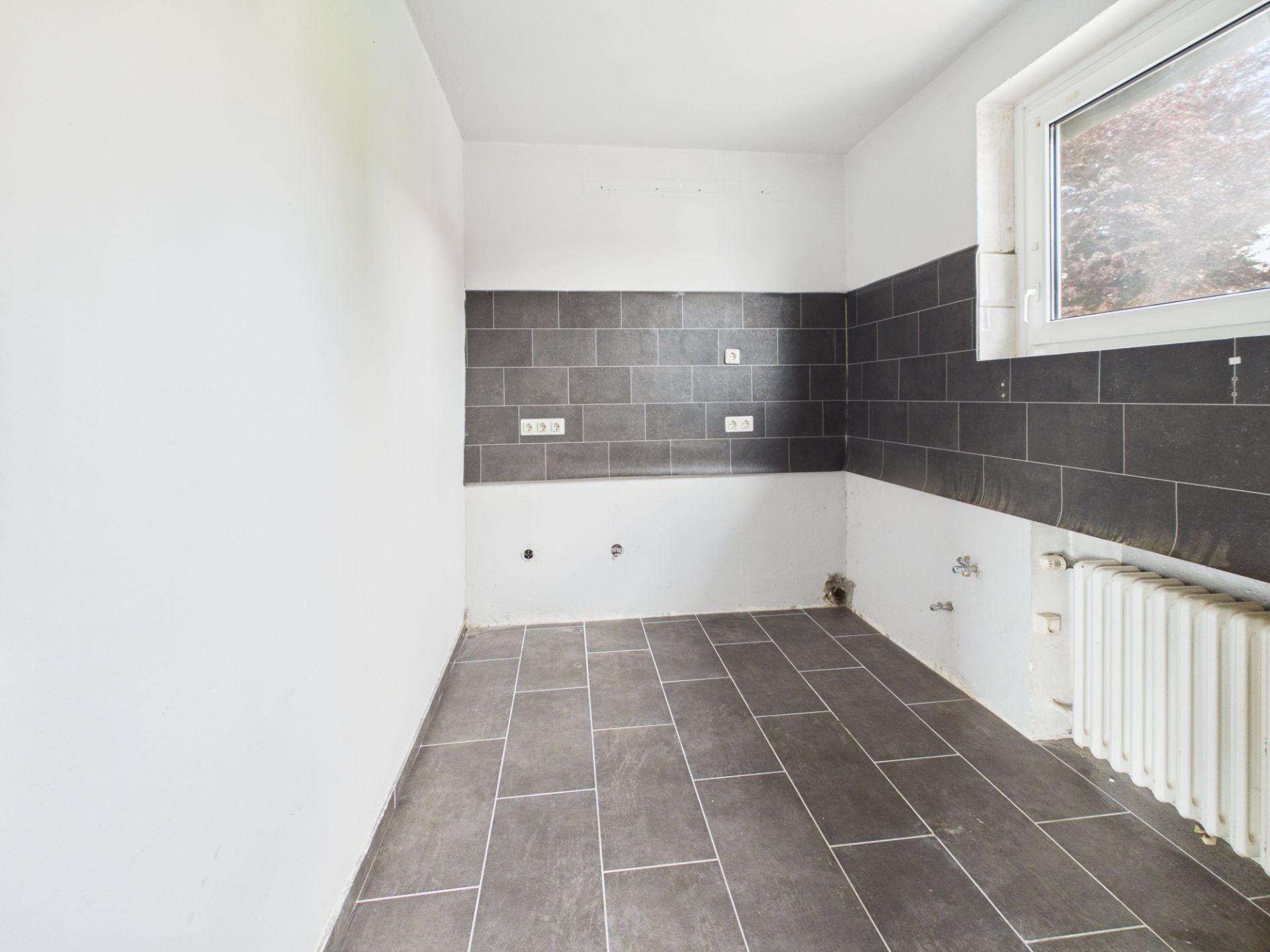 Leerer Raum mit grauem Fliesenboden, weißer Wand und Fenster, schwarze Wandfliesen, Heizkörper rechts.