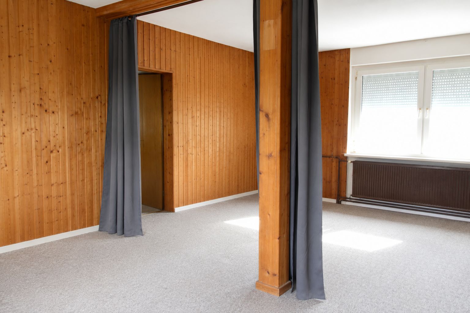 Leerstehender Raum mit Holzverkleidung, grauen Vorhängen und großem Fenster.