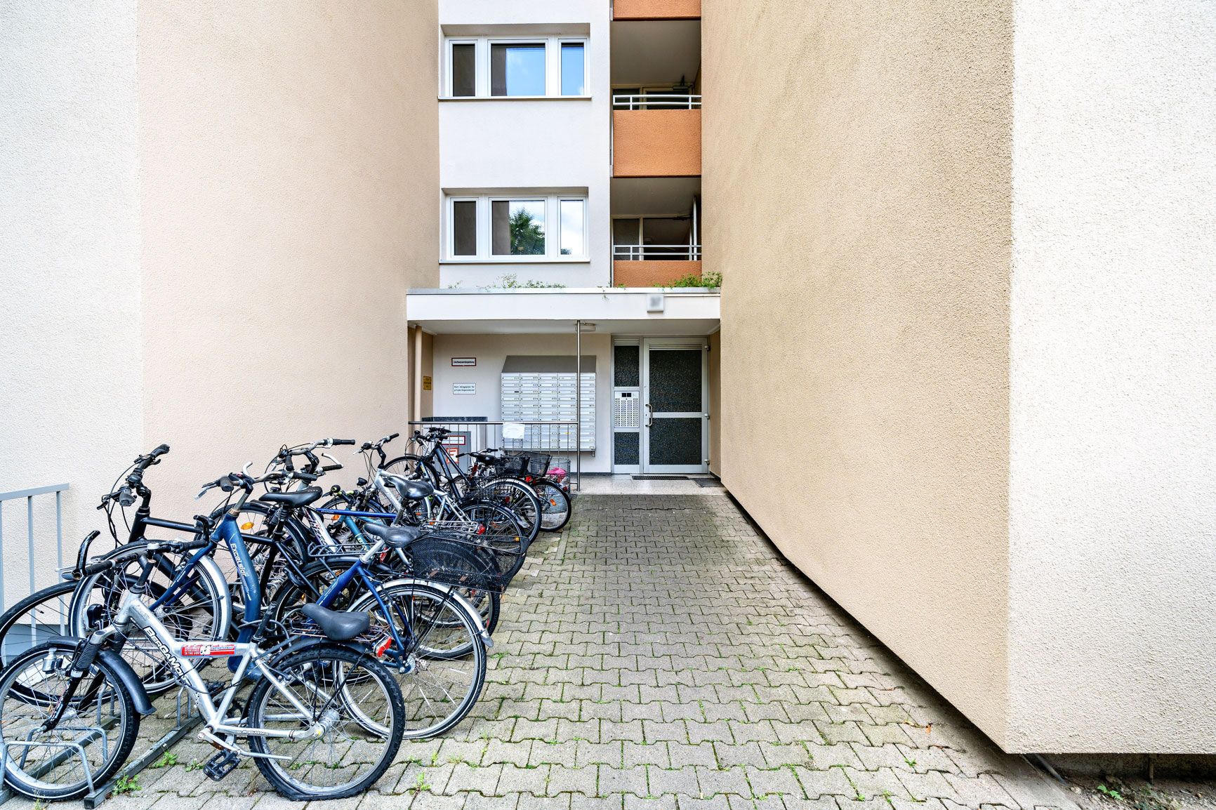 Eingang eines Wohnhauses mit mehreren abgestellten Fahrrädern auf gepflastertem Weg.