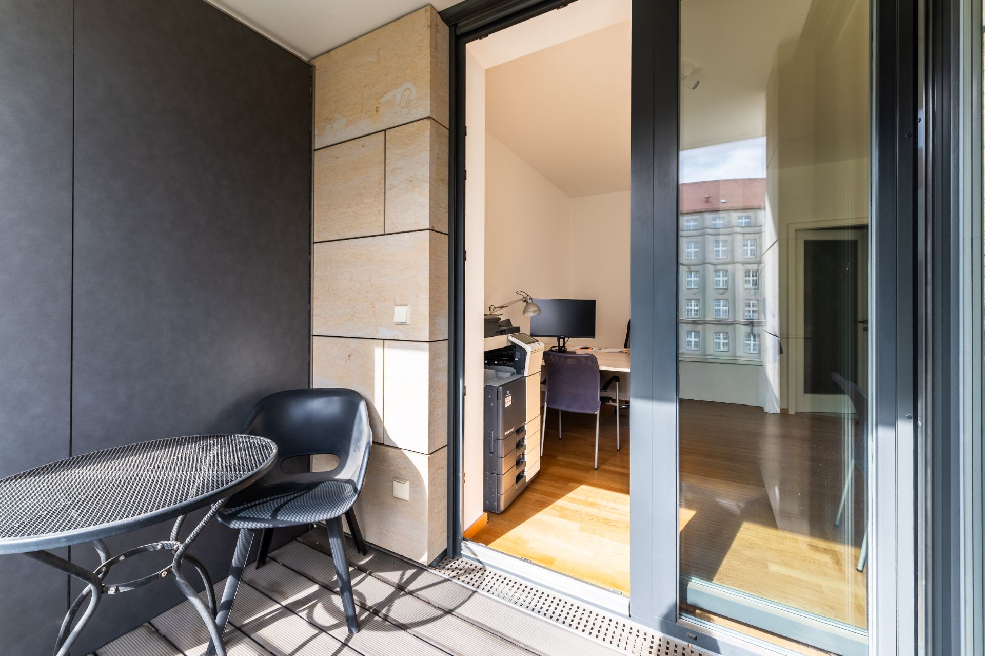 Balkon mit zwei schwarzen Stühlen und Tisch, Zugang zu einem hellen Arbeitszimmer mit Schreibtisch und Monitor.