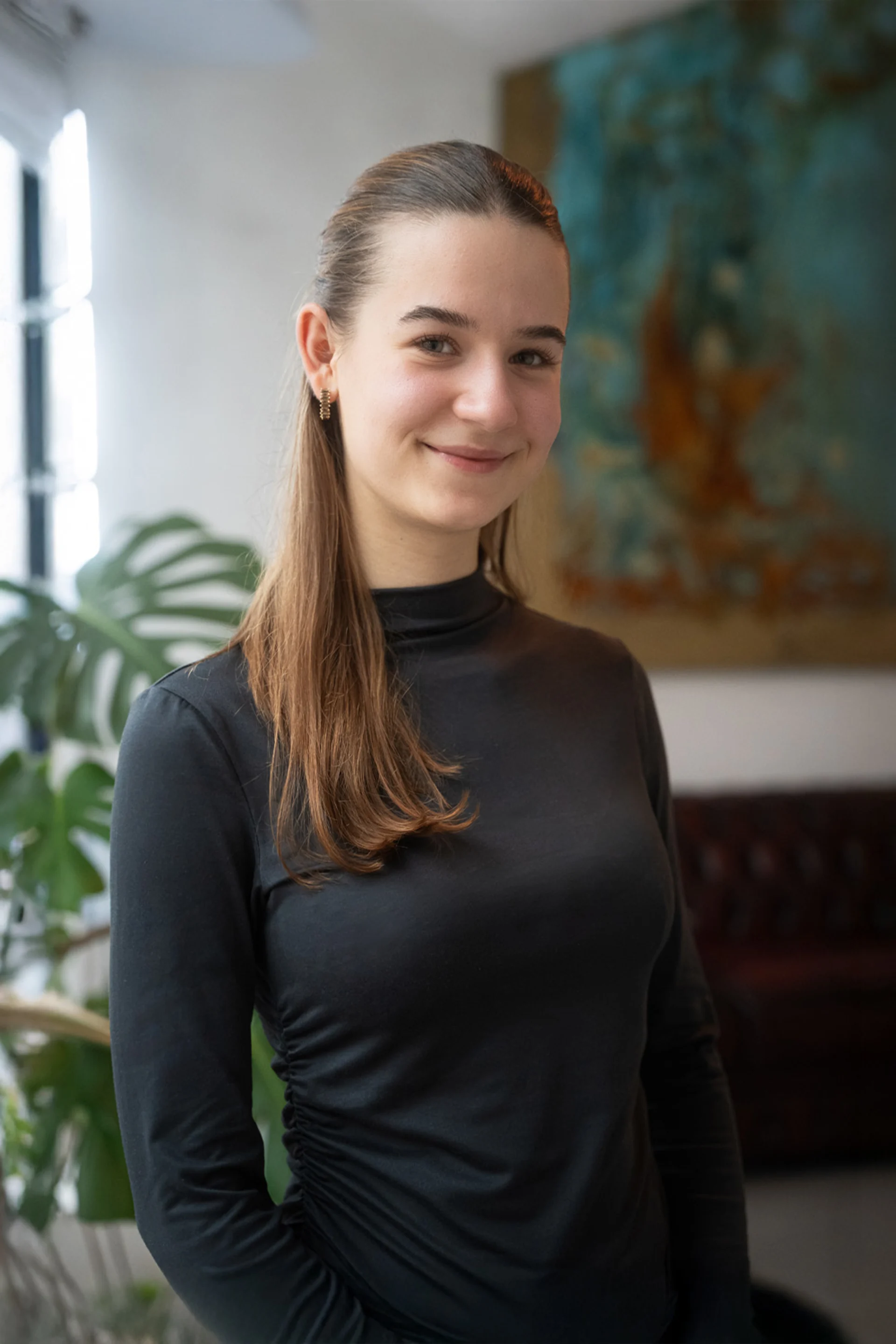 Junge Frau mit langen Haaren steht lächelnd in einem Wohnzimmer mit Pflanze und Sofaecke.