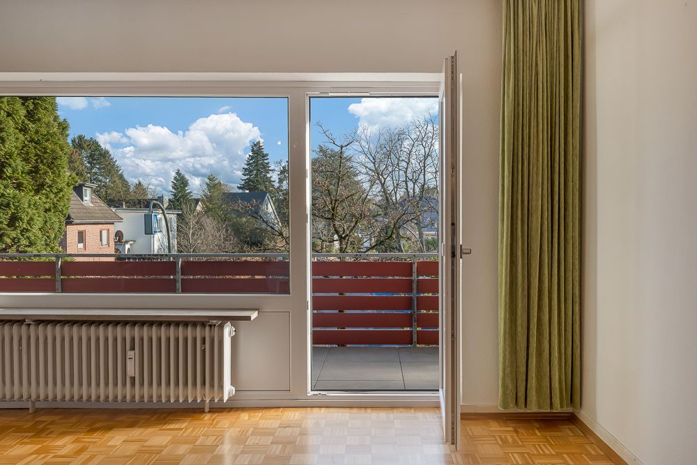 Blick aus einem Zimmer mit Parkettboden auf einen Balkon und grüne Aussicht.