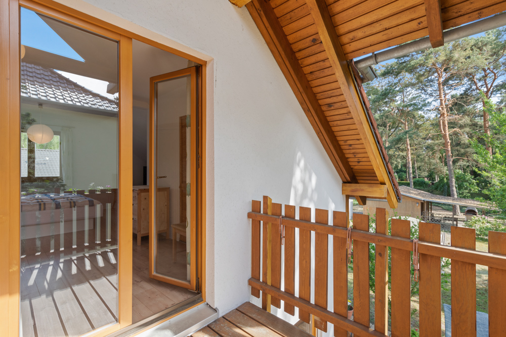 Kleiner Balkon mit Holzzaun und Blick auf Bäume, angrenzend an ein Zimmer mit Glastür.