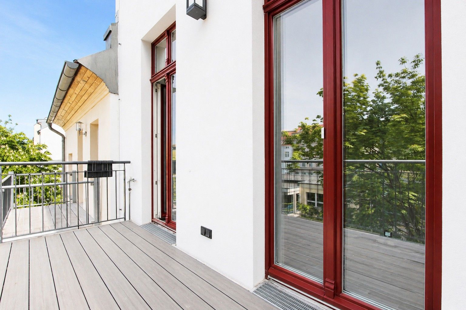 Helle Terrasse mit Holzgeländer und großen Glastüren, Bäume im Hintergrund.