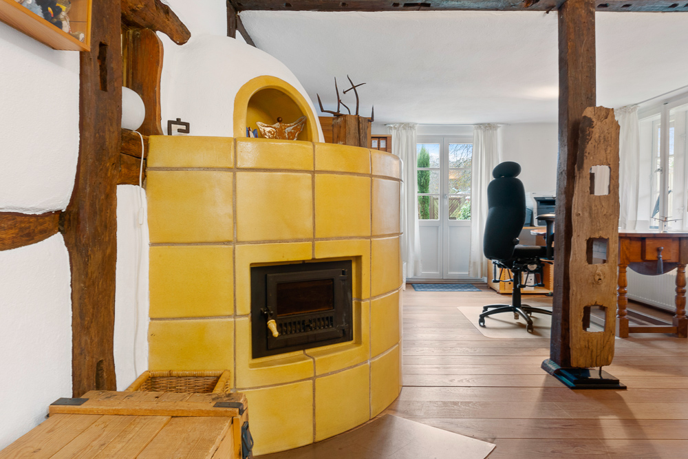 Gelber Kachelofen steht in einem hellen Raum mit Schreibtisch, Stuhl und Holzbalken.