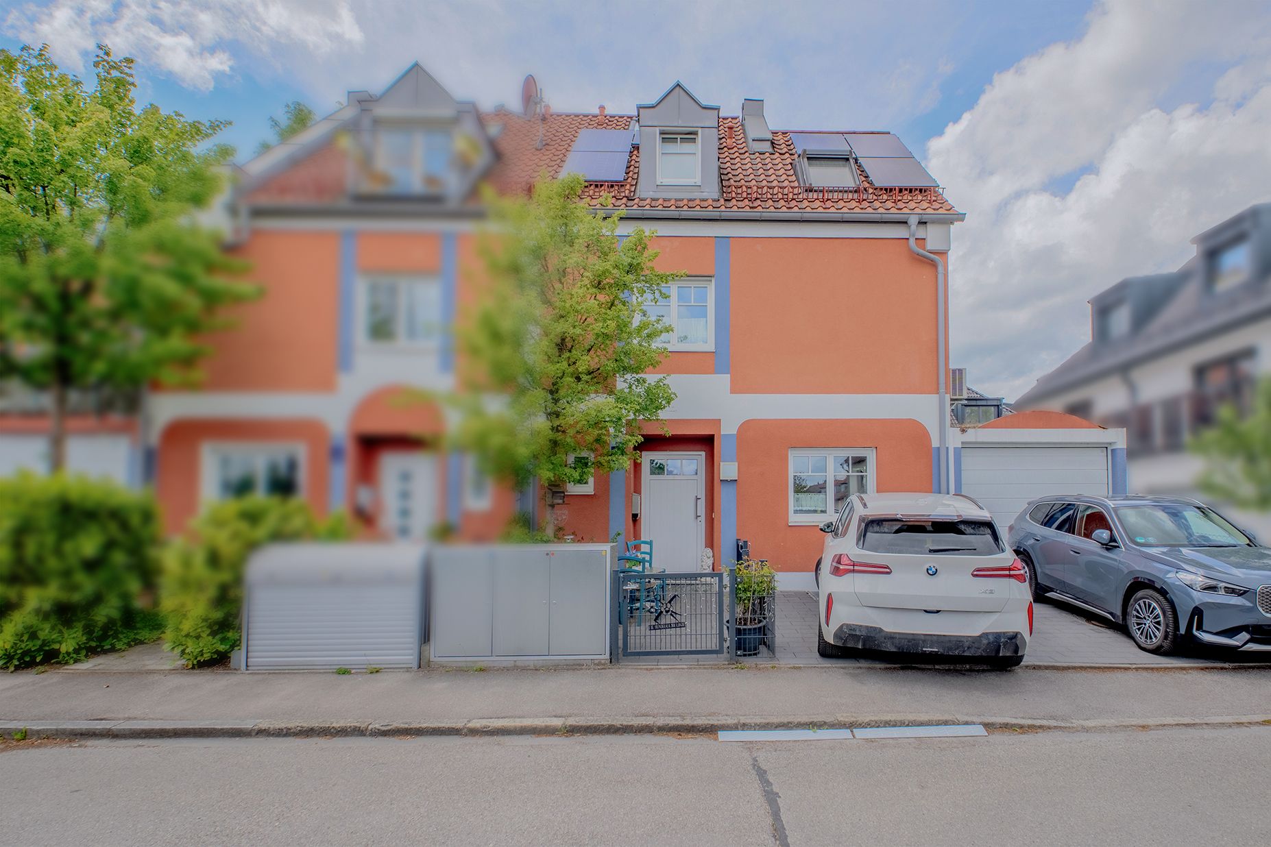 Reihenhaus mit orangefarbener Fassade, zwei geparkten Autos und Bäumen davor.