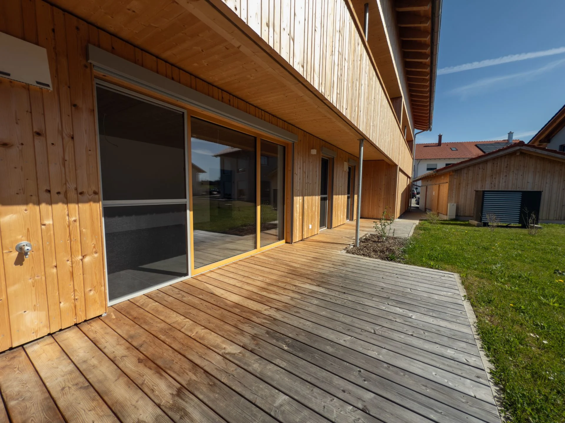 Holzterrasse mit großem Fenster und Rasenfläche neben einem modernen Wohnhaus.