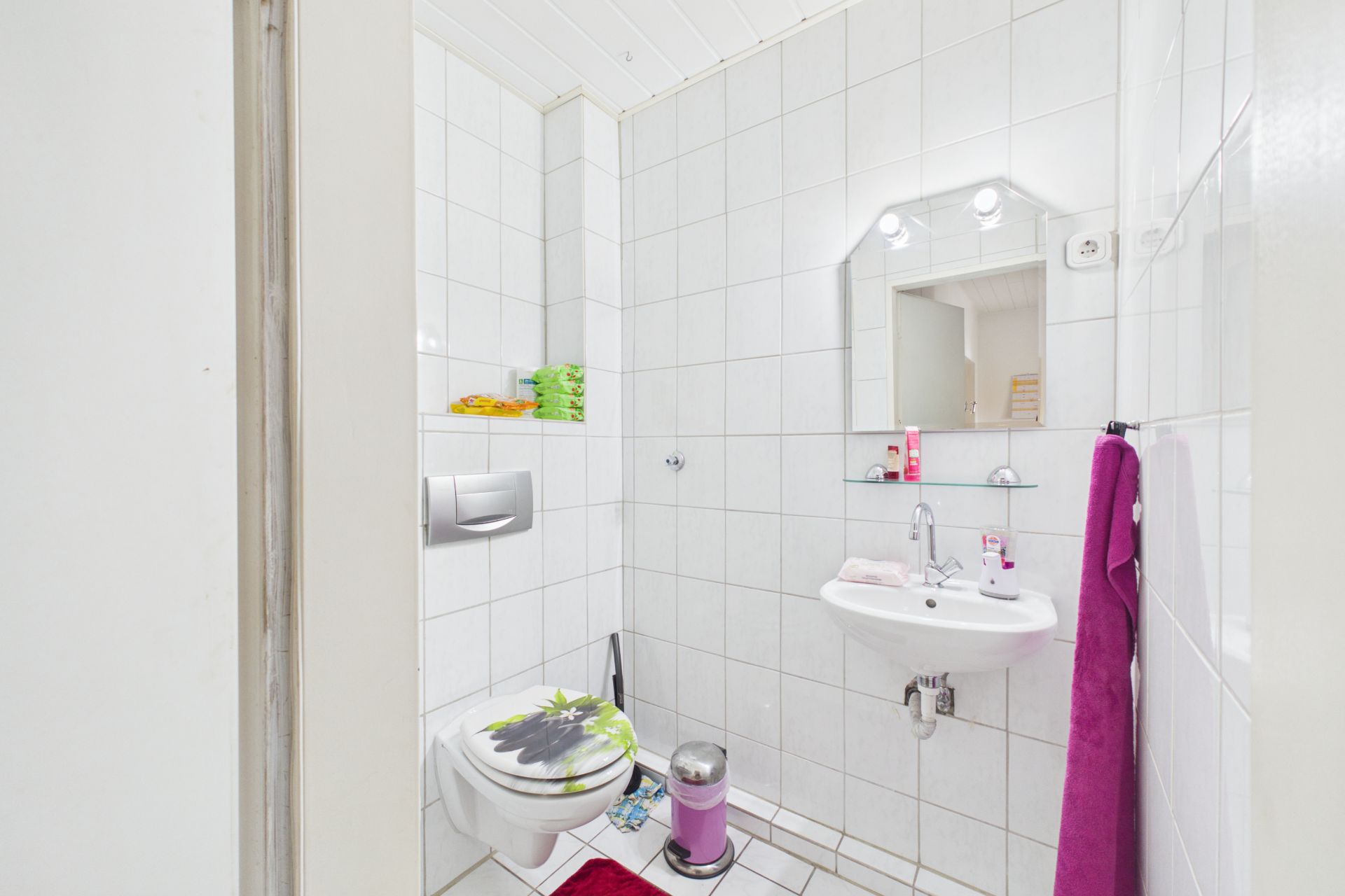 Kleines Bad mit WC, Waschbecken, Spiegel und weißen Fliesen, lila Handtuch und Mülleimer.