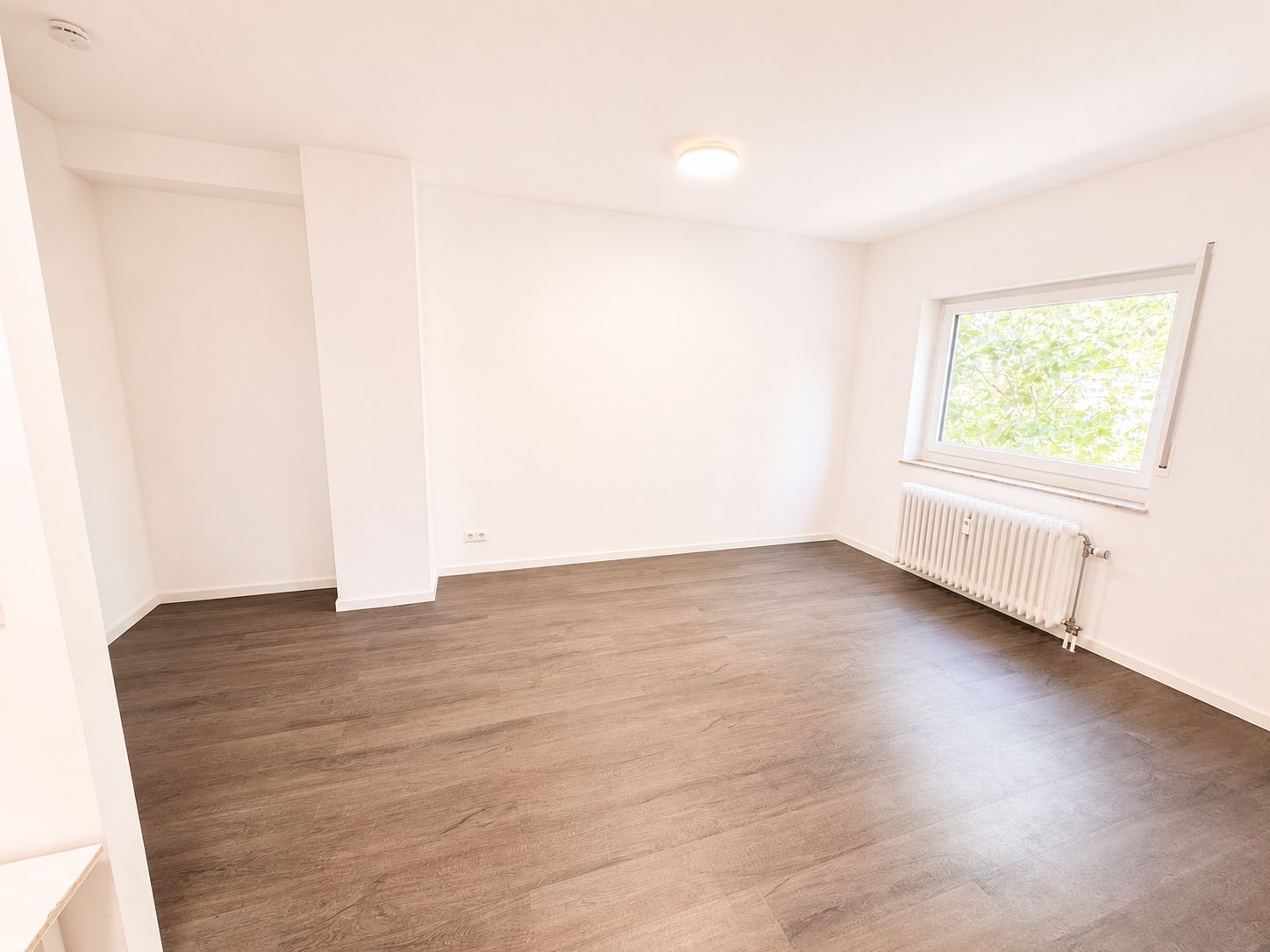 Helles leeres Zimmer mit weißer Wand, großem Fenster, Heizkörper und dunklem Holzboden.