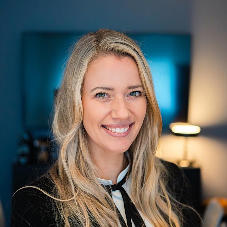 Junge Frau mit langen blonden Haaren lächelt in einem modern eingerichteten Raum.