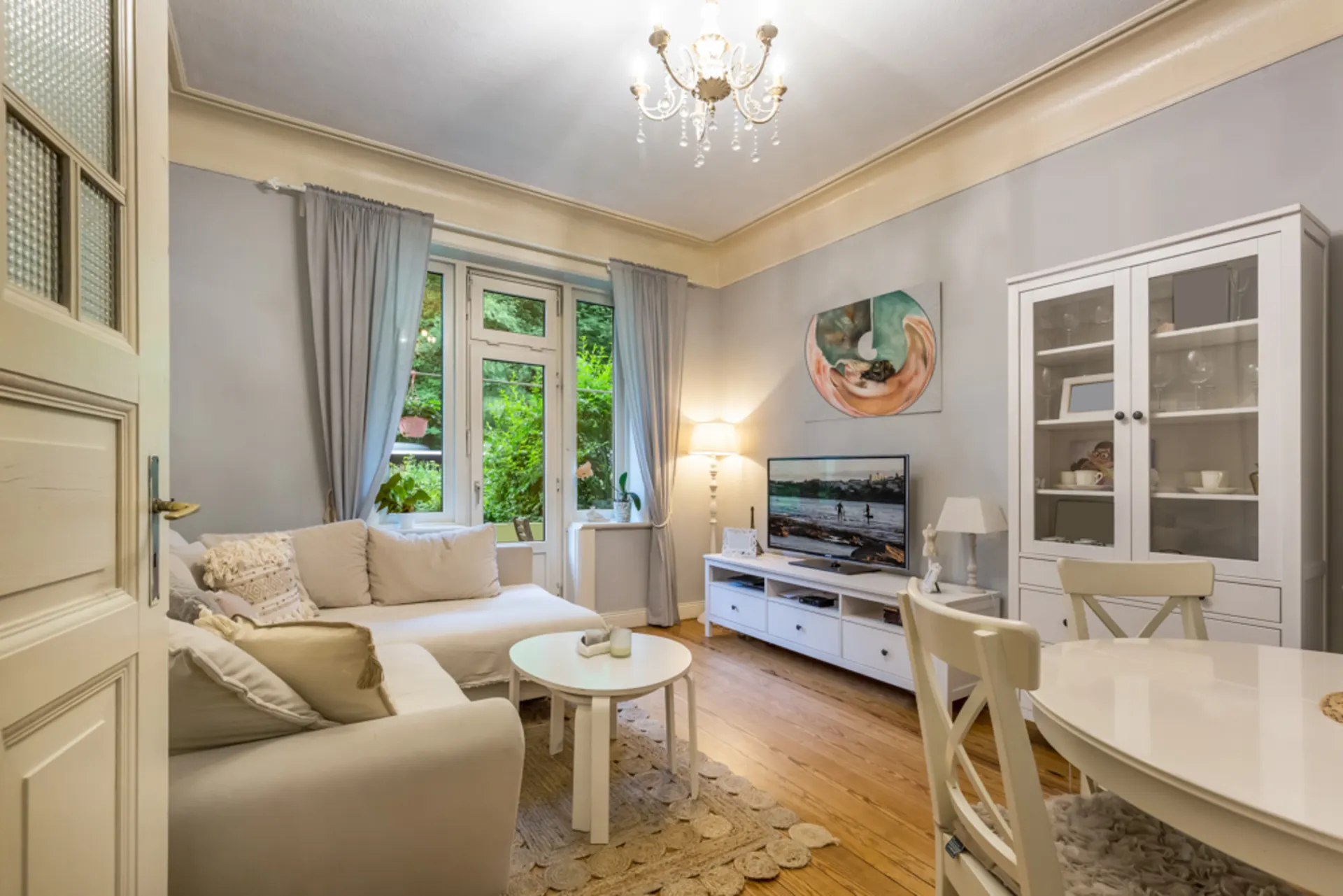 Helles Wohnzimmer mit Sofa, TV, Esstisch, Vitrine und großen Fenstern mit Vorhängen.