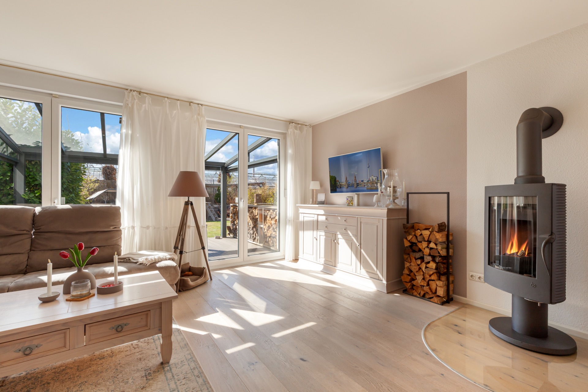 Helles Wohnzimmer mit Kamin, Sofa, Couchtisch, TV, Holzstapel und großen Fenstern zum Garten.