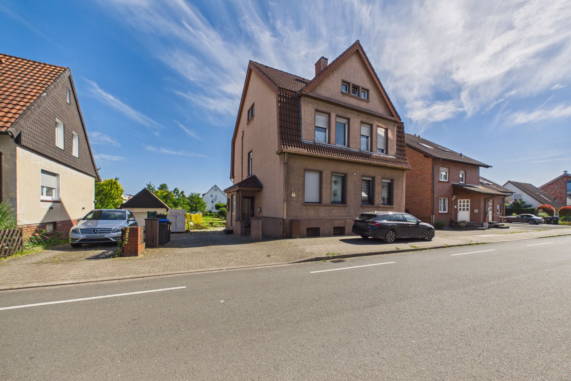 Einfamilienhaus mit braunem Dach an einer ruhigen Straße, zwei Autos parken davor.