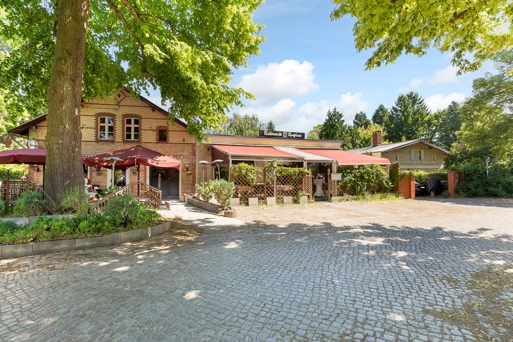 Backsteinhaus mit Terrasse, großen Bäumen und Kopfsteinpflaster im Vordergrund.