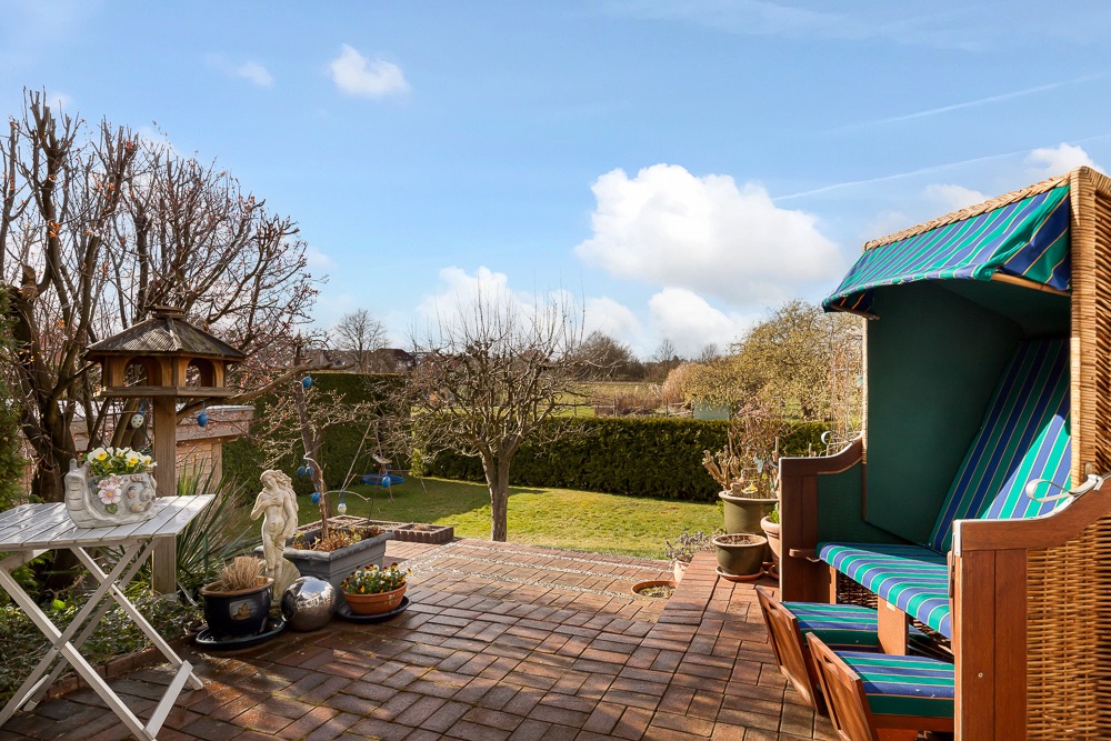 Gepflasterte Terrasse mit Strandkorb, Gartenmöbeln, Pflanzen und Blick auf einen grünen Garten.