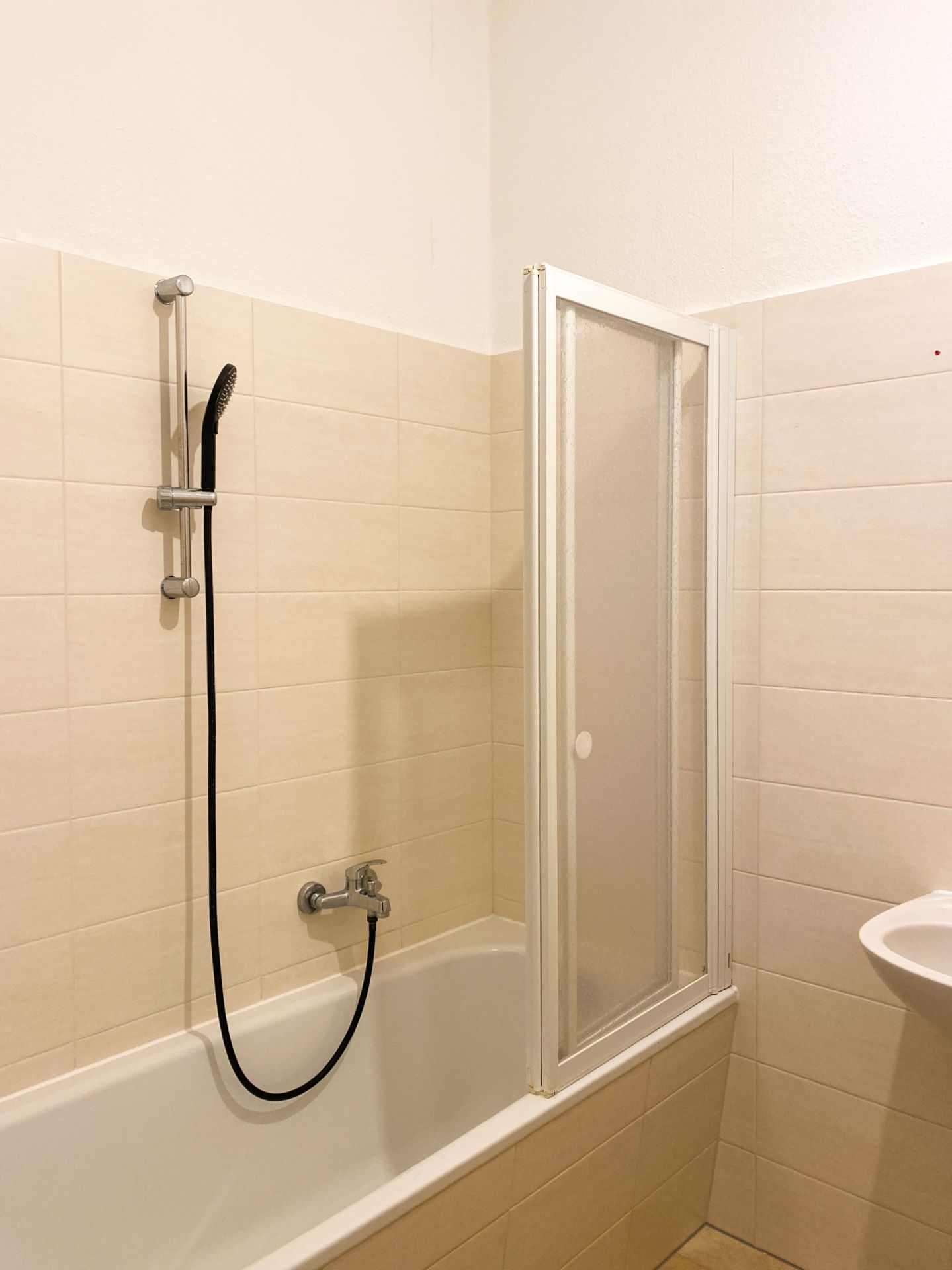 Badewanne mit Duschbrause und Glas-Duschabtrennung, helle Fliesen an der Wand.