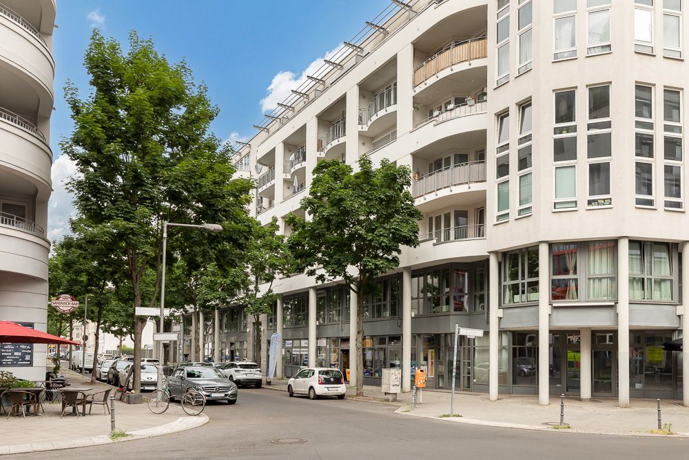Moderne Wohngebäude mit Balkonen an einer Straßenecke, Bäume und geparkte Autos sichtbar.