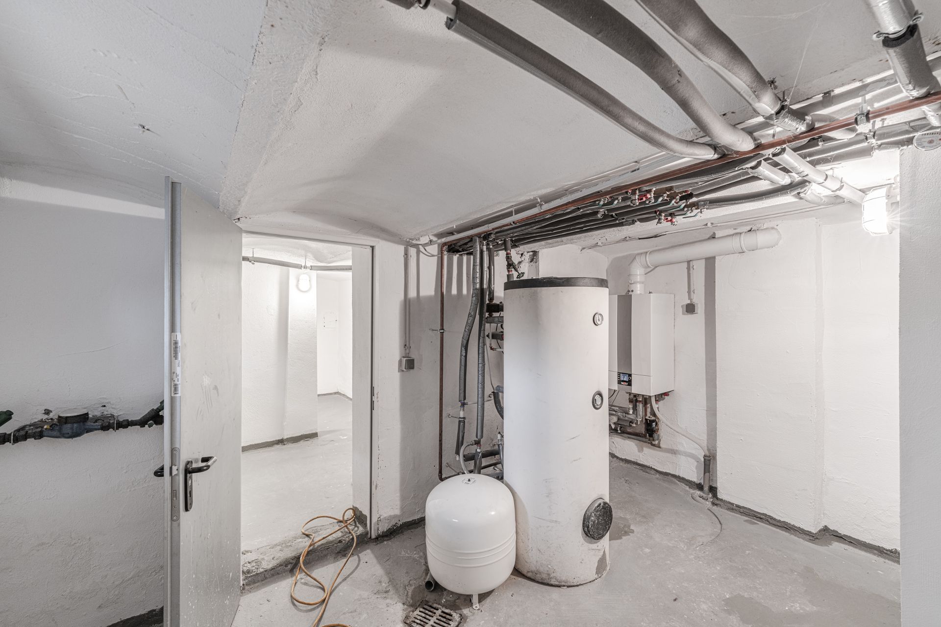 Heller Kellerraum mit Heizungsanlage, Rohren und offen stehender Tür.